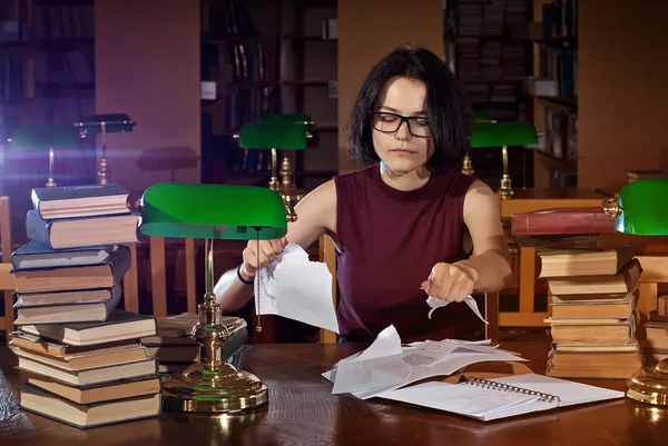 brunette in glasses at the table with books. Library with green lights on the tables. An old library with brown furniture.