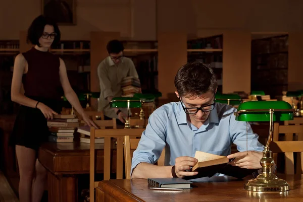 guy in glasses at the table with books. Library with green lights on the tables. An old library with brown furniture.