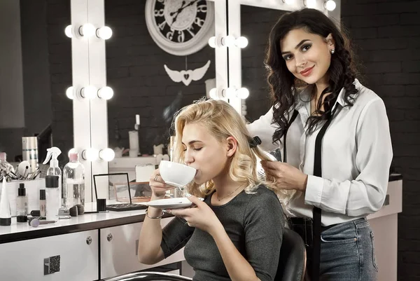 这个女孩在美容院喝咖啡 向导会为客户端制作发型 金发和黑发背景的镜子与背光 精细的客户服务 — 图库照片