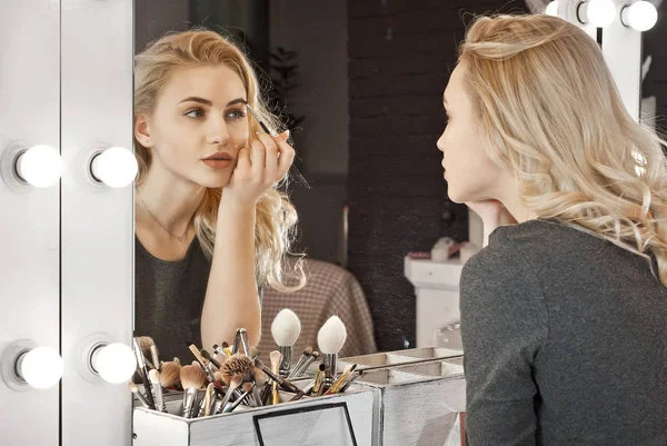 The girl makes a makeover near the mirror with a backlight. A pretty blonde in a beauty salon.