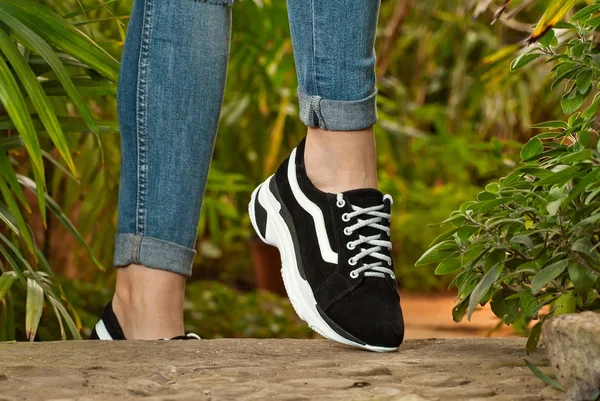 Female legs close-up. Sports sneakers on asphalt against green plants. Shoes in a tropical climate arranger.