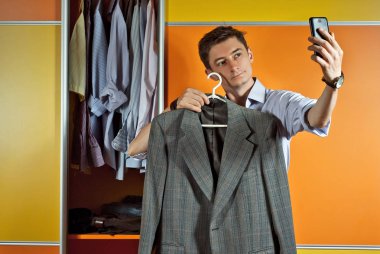 Çizgili gömlekli bir adam dolabın arka planında kıyafet dener. Sarı ve turuncu gardırop. Adam telefonda takım elbiseli sephi yapıyor..