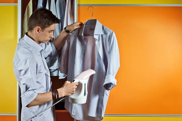 A man in a striped shirt tries clothes. Yellow and orange wardrobe. The guy strokes his shirt with a steam iron. Concept of clothing selection and fitting. Preparation and care of clothes.