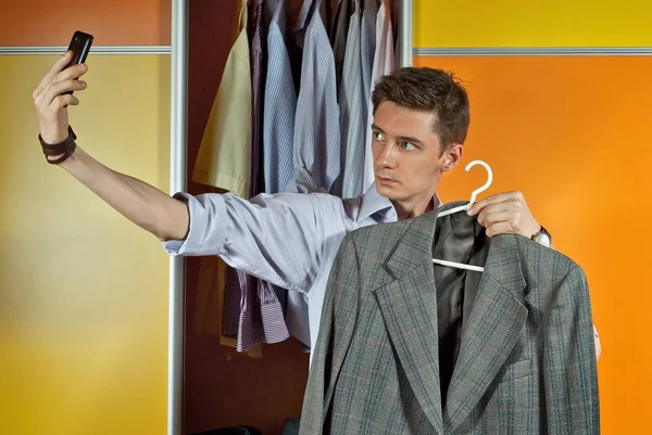 A man in a striped shirt tries clothes on the background of the closet. Yellow and orange wardrobe. The guy makes a sephi with a suit on the phone.