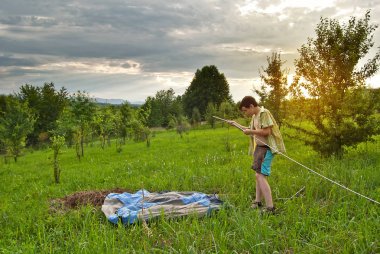 Erkekler çimlerin üzerine çadır kurarlar. Arka planda orman ve dağlar. Güneş geliyor ve gökyüzü sarı.. 