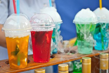 Çok renkli meyveli içecekler. Festivalde plastik bardaklarda alkolsüz içkiler. Meyve suyu ve sebzeli kokteyl. Beyaz duman kokteyllerle buharlanıyor..