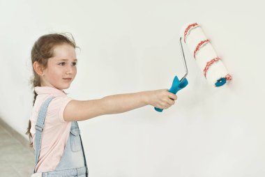 Küçük kız mavi tulumla tamir atıyor. Çocuk duvarları boyar. Beyaz duvar arka plan üzerinde elinde rulo ile Kız.