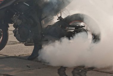 Duman tekerleklerin altından çıkıyor. Motosiklet tekerleği yakın plan. Lastiklerin asfalta sürtünmesinden dolayı duman. Sürücü motosiklet üzerindeki numarayı yapmaya hazırlanıyor. Yolda yanmış lastik..