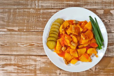 Tahta bir masada beyaz bir tabakta patates. Et ve soğanlı dilimlenmiş patates. Portakal soslu haşlanmış patates. Eski pembe bir tahtanın arka planında mutfak gereçleri var. Tasarım için alanı kopyala.