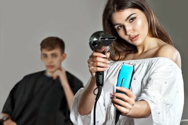 Elinde saç kurutma makinesi ve makasıyla kız kuaförü. Arka plandaki adam saçını kestirmek için bekliyor. Kadın müşterinin saçını kesmeye hazırlanıyor. Saç kurutma makinesi ve tıraş makinesi olan bir kuaför..