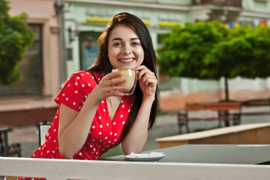 Bir sokak kafesinin masasında oturan mutlu kız. Kadın beyaz noktalı elbiseyle kırmızı kahve içiyor..