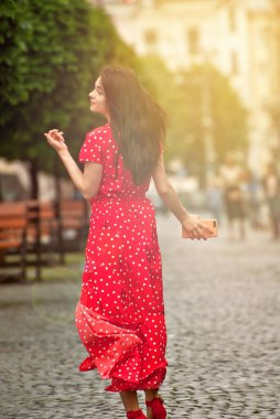 Mutlu kız kaldırım taşlarıyla sokakta yürüyor. Beyaz lekeli kırmızı elbiseli bir kadın. Elbiseler ve saçlar rüzgar tarafından uçurulur. Bulanık şehir sokağında arka planda bir kız.