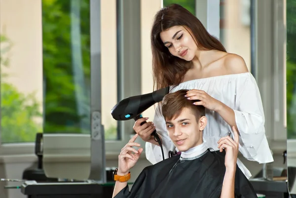 The girl makes a hairstyle to the guy. Hairdresser in a beauty salon with a hair dryer in his hands.