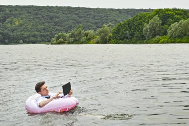Pembe şişme dairede tişörtlü bir adam. Dizüstü bilgisayarı olan adam nehirde yüzüyor. Tatil, taşeronluk ve serbest çalışma kavramı. İş adamının yanında metin ve kopyalama alanı.