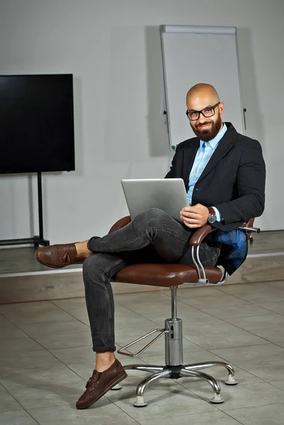 Businessman in a blue shirt sitting in a brown leather chair. Man in a jacket with a laptop in glasses. Bearded office worker at work. Monitor and drawing board in the background.