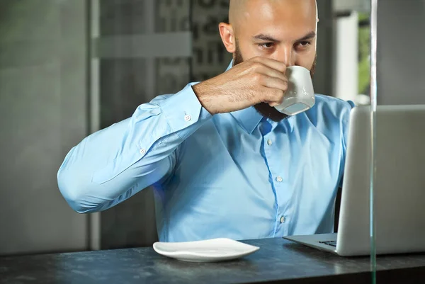 Businessman in a blue shirt sitting at a table with a laptop. Bearded man working in the office. Office worker drinking coffee from a white mug.