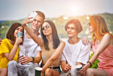 Arkadaşları şehrin arka planında kahve içiyor. Erkekler ve kızlar gülümsüyor. Meydanda güneşli bir günde kaldırımda oturan gençler. Gençler selfie çekiyor..