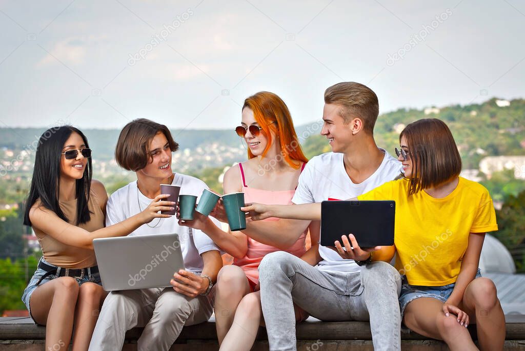 Friends sitting on the curb with a laptop in the summer. Teenagers with disposable paper cups drinking coffee. Online learning concept.