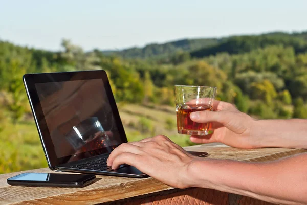 Man working on laptop. Work on nature background. Sunny summer day. The concept of remote work and freelance. Hands and laptop close up. A glass of whiskey or tea in hand.