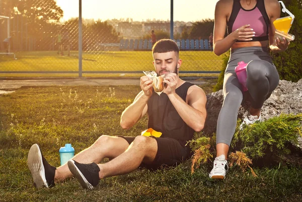 Man and woman eating hamburger after workout. Athletes rest together. Couple on a sunset background.