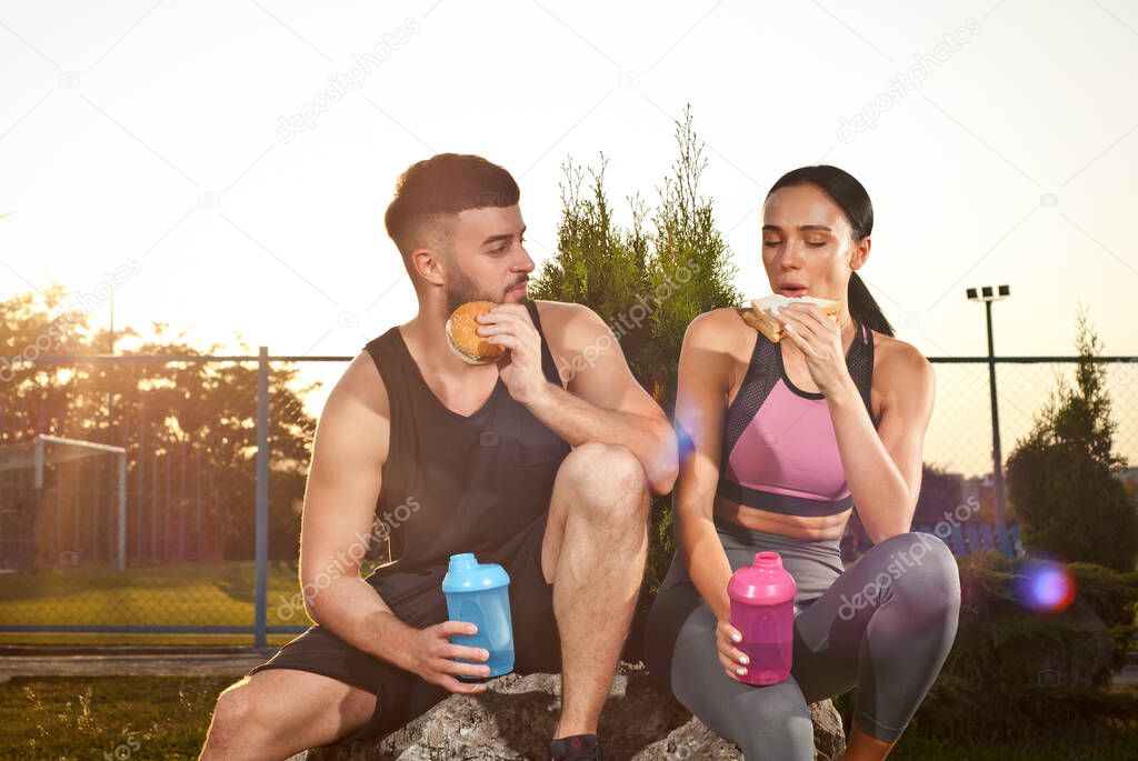 Man and woman eating hamburger after workout. Athletes rest together. Couple with water bottles on sunset background.