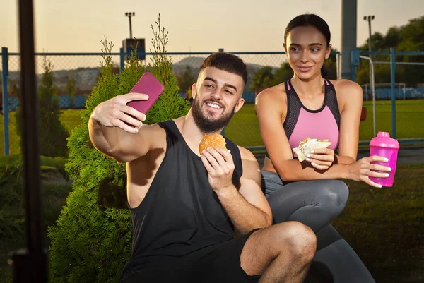 Man and woman eating hamburger after workout. Athletes rest together. Couple with water bottles on sunset background. Friends take selfies.