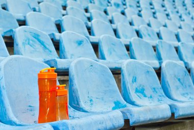 Stadyumda bir sürü boş koltuk var. Koltukta portakal suyu şişesi var. Sporcular için iç. Plastik koltuklar sıraya dizildi. Kirli ve çizik mavi fan koltukları.