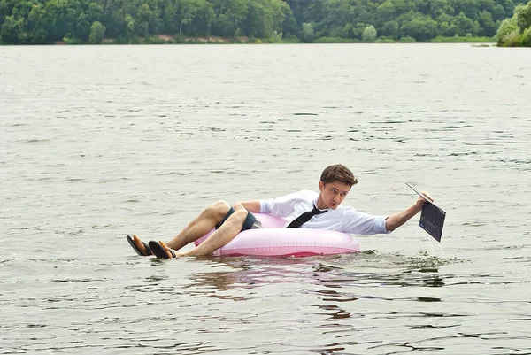 A man in a shirt on a pink inflatable circle. Guy with a laptop swims in the river. The concept of work during vacation, outsourcing and freelance. The man drowned the computer. Mishap.