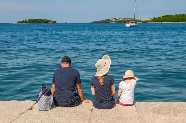 rear view of the family sitting on a stone pier on a summer day. The family admires the beautiful sea view from the shore