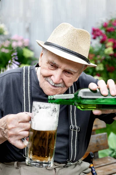 elderly man in a hat drinks beer outside a pub on a bench in the garden. The old man pours beer into a glass. Close up.