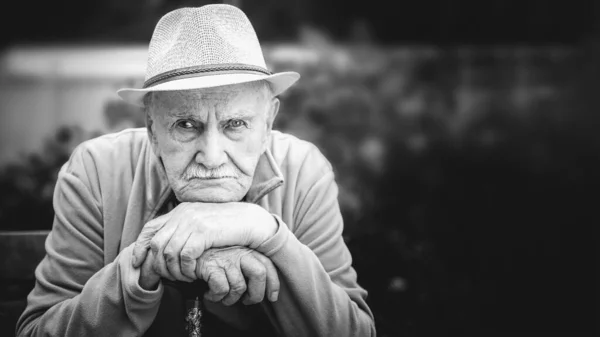 sad, thoughtful, serious old man in a hat is sitting in an open-air garden. the concept of loneliness and lonely old age. Black and white portrait. Banner, copy space.