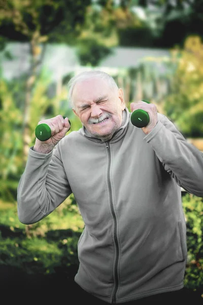 An old man trains his muscles with dumbbells in the Park after a home quarantine at the time of the pandemic. Active old age and healthy lifestyle in retirement