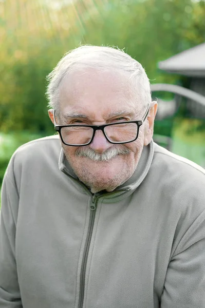 cheerful gray-haired old man looks at the interlocutor and smiles. The old man can\'t see very well without his glasses. Portrait of a cheerful old man with glasses close-up.