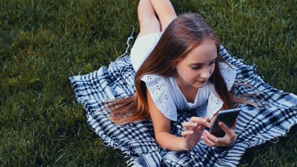 Счастливая девушка печатает смс по телефону лежит на траве в парке. Цветовой сорт — стоковое видео