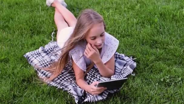 Happy girl using tablet pc lies on grass in the park — Stock Video