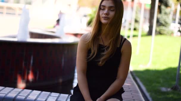Jonge blonde vrouw zittend op een bankje met fontein op achtergrond — Stockvideo