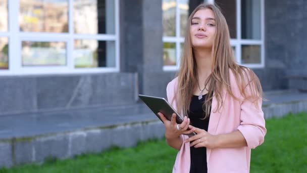 Ung blondine står med en tablet i hænderne. Kvinde bruger trådløst internet på gaden. Mobil kommunikationskoncept . – Stock-video