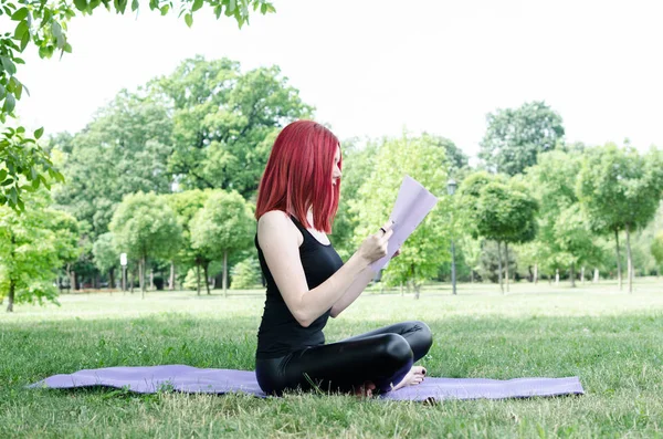 Young woman or girl reading and teaching yoga in nature. Woman holding paper and document or book and sitting on grass in nature