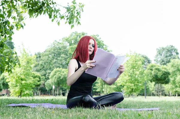Young woman or girl reading and teaching yoga in nature. Woman holding paper and document or book and sitting on grass in nature