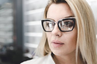 Beyaz üniforma giyen karizmatik, kendine güvenen düşünülmeli. Kadın moda lens takmış. Doktor şık ve modern optik gözlük şeffaf ayaklıklı önünde duruyor.