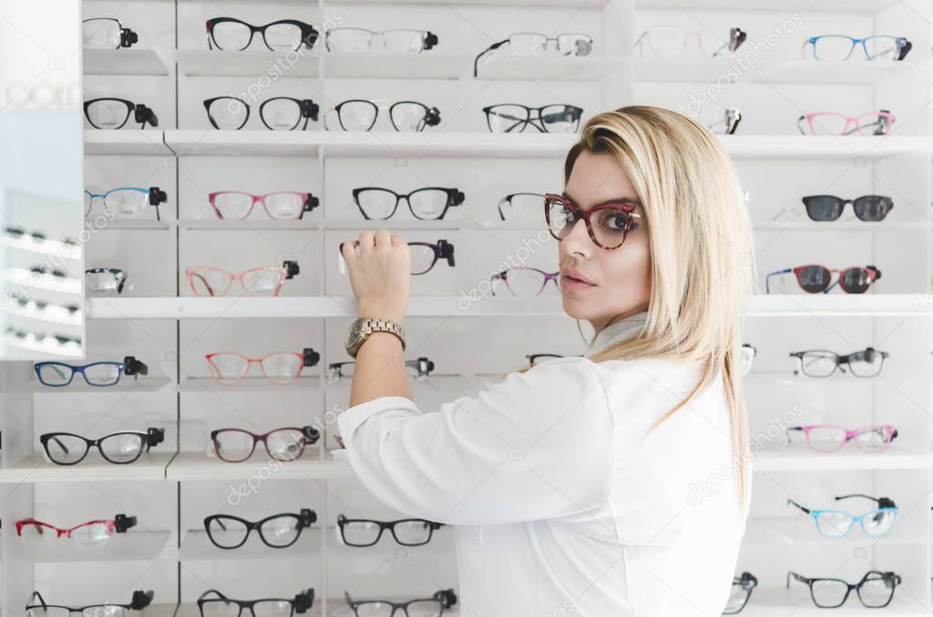 Charismatic, confident oculist wearing white uniform. Woman wearing fashionable lenses. Doctor is standing in front of transparent stand with stylish, modern optical eyeglasses.