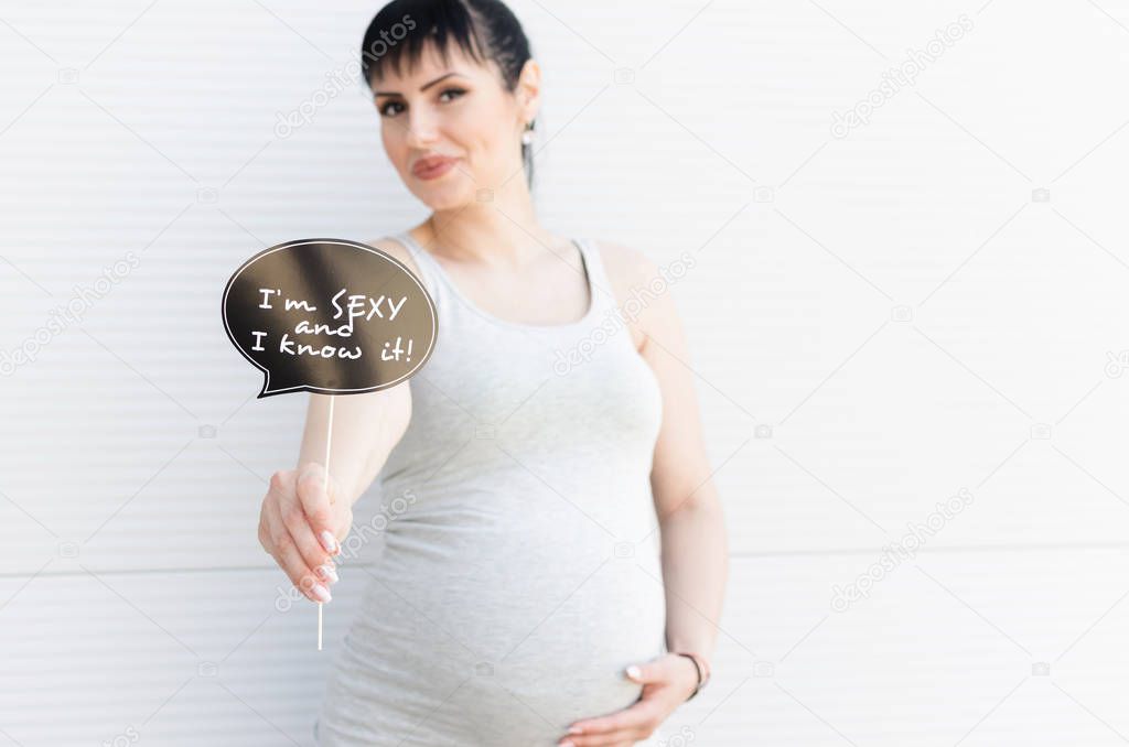 Beautiful young pregnant woman holds photo booth prop with funny quote.