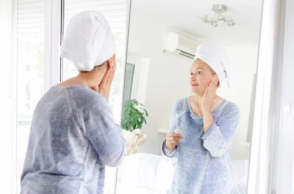 Smuk Senior Kvinde Der Anvender Age Creme Foran Spejlet Derhjemme - Stock-foto