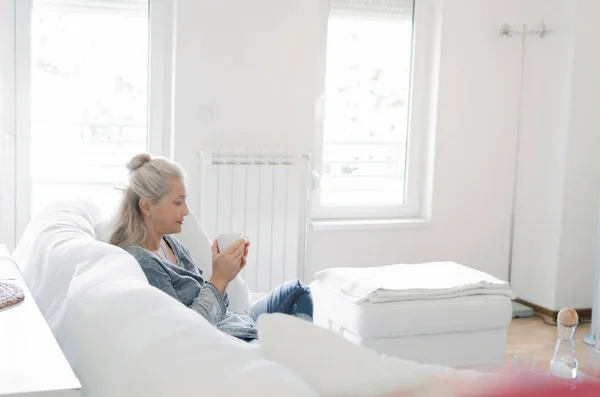 Senior woman sitting on couch in livingroom holding cup in hands, space for text.