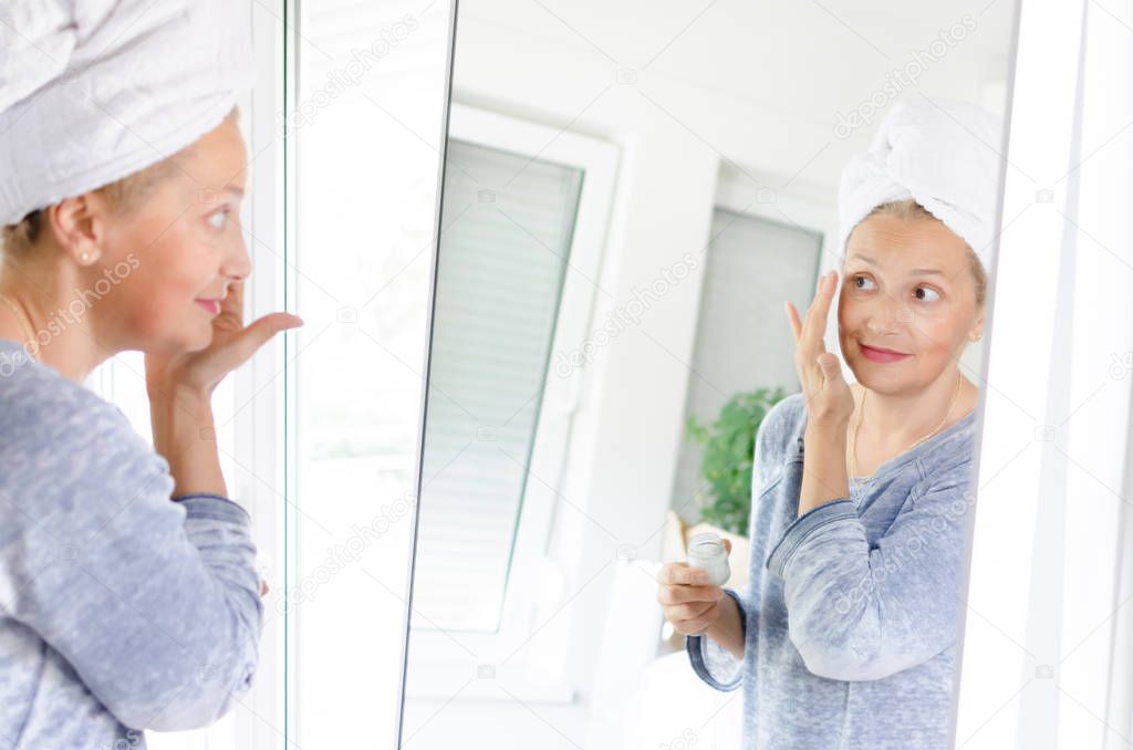 Beautiful adult woman applying face cream in front of mirror at home.