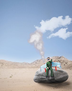 Jetpack kanatları ile çölde kaya üzerinde oturan havacı kostümlü Yaratıcı küçük çocuk