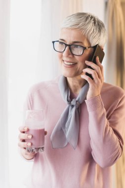 Modern kısa gri saç modeli ve gözlük pencere tarafından ayakta ve cep telefonu konuşurken güzel olgun kadın 