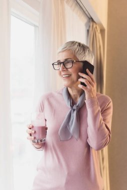 Modern kısa gri saç modeli ve gözlük pencere tarafından ayakta ve cep telefonu konuşurken güzel olgun kadın 