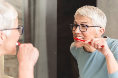 Beautiful senior woman washing her teeth and looking at her reflection in mirror clipart