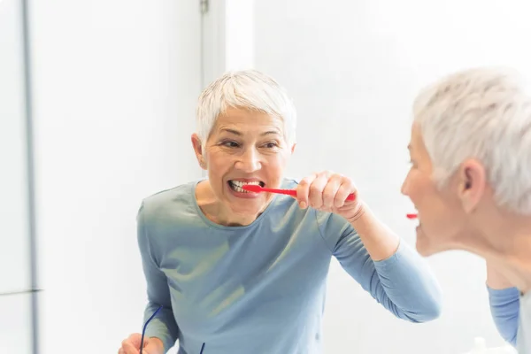 Mirror reflection of mature woman washing her teeth with red toothbrush in bright bathroom